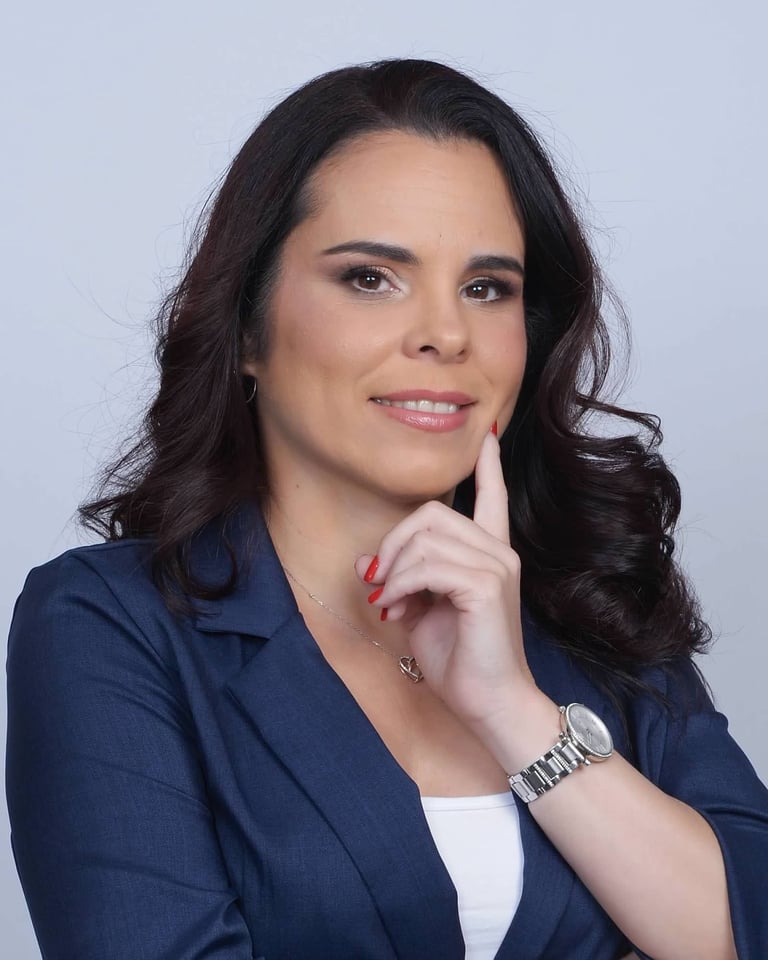 Professional headshot of a woman with long dark hair wearing a dark blue blazer and white top, posed with her hand to her chin