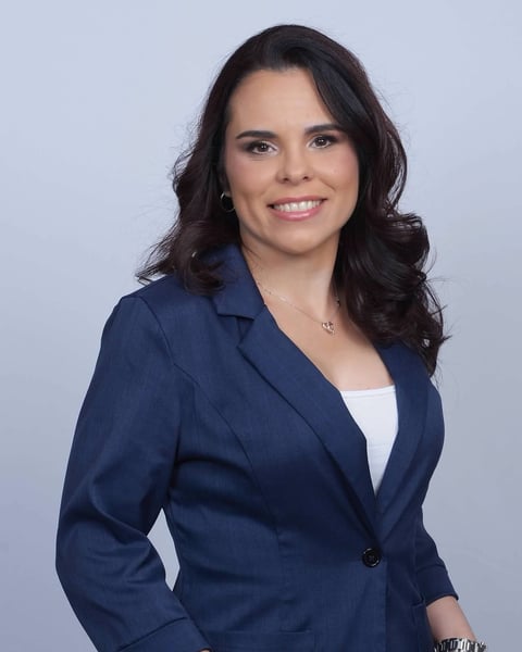 Professional headshot of a woman with long dark hair wearing a navy blue blazer and white top, smiling against a light blue background